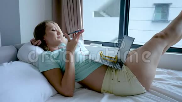 成年女性穿着白色床单和小玩意躺在现代公寓里有大窗户和黑暗的框架并倾听视频的预览图