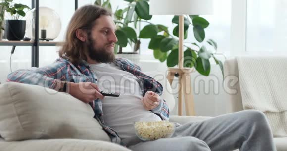 带爆米花和电视遥控器的男人坐在家里的沙发上视频的预览图
