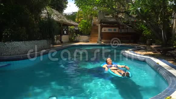 阳光明媚的一天年轻女子躺在游泳池里的水床垫上视频的预览图