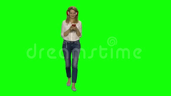 金发女人从容地走在大红耳机里在绿色屏幕上的手机上选择音乐铬键4k视频的预览图