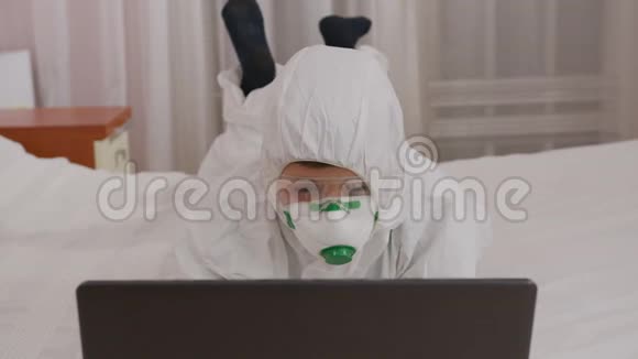 一个穿着防护服和呼吸器的男孩在家里的电脑上被隔离在流行病期间视频的预览图