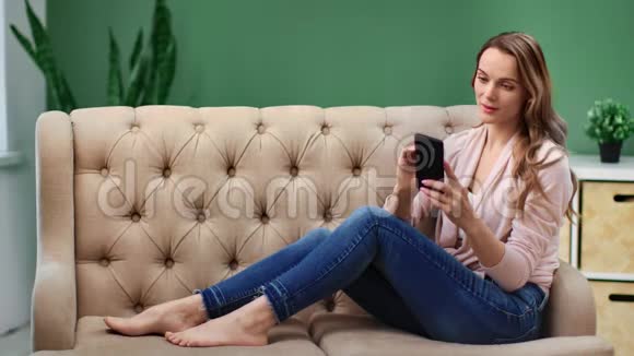 微笑的女性聊天使用智能手机坐在舒适的沙发上红色相机宽镜头视频的预览图