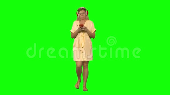 晒黑的金发女人正平静地戴着红色的大耳机走着在绿色屏幕上的手机上选择音乐正面观点视频的预览图