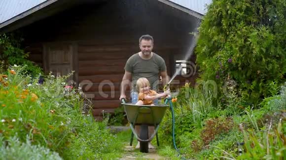 在温暖的阳光明媚的日子里快乐的小男孩坐在爸爸推着的独轮车里玩从茅屋里给植物浇水的小孩视频的预览图