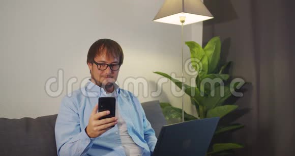 快乐的年轻人使用笔记本电脑和智能手机工作而坐在沙发在家里视频的预览图