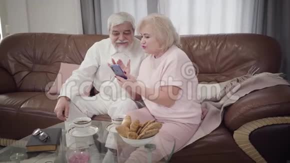 成熟的白种人男女在家使用智能手机的肖像现代退休人员坐在室内沙发上聊天视频的预览图