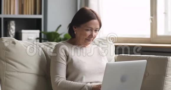 中年老年妇女用坐在沙发上的笔记本电脑打字视频的预览图