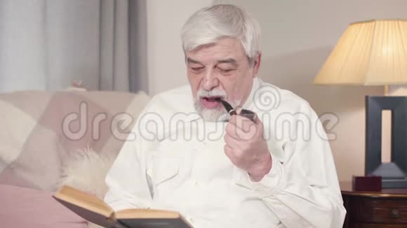 白发苍苍的白人男子在家抽烟斗和看书的肖像成熟严肃的男人度过安静的夜晚视频的预览图