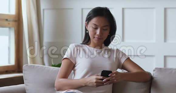 在沙发上使用智能手机应用程序微笑着的千禧亚洲女性视频的预览图