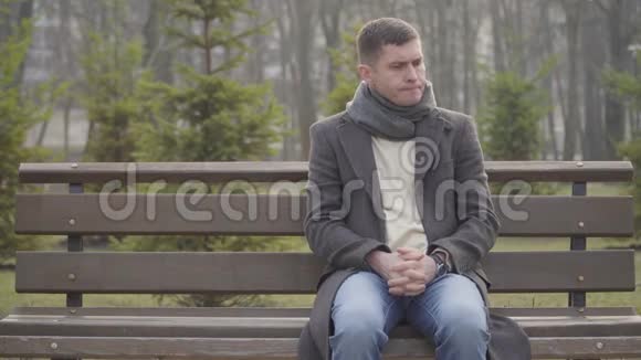 紧张的白人年轻人坐在城市公园的长凳上看着时间不耐烦的黑发男人的肖像视频的预览图