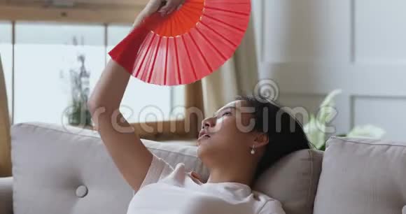 恼火过热的亚裔女子挥舞扇子坐在室内沙发上视频的预览图