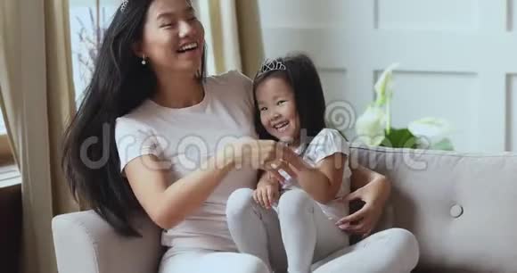 活泼开朗的亚洲妈妈爱挠痒痒的女儿抱着她坐在沙发上视频的预览图