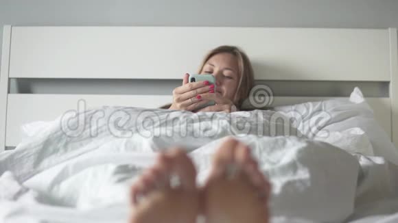 一个年轻的女人早上躺在床上拿着手机视频的预览图
