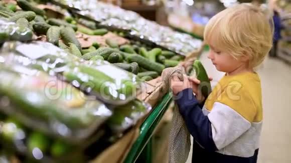 可爱的小男孩选择新鲜的有机黄瓜并放入网状购物袋在食品商店或超市零废物概念视频的预览图