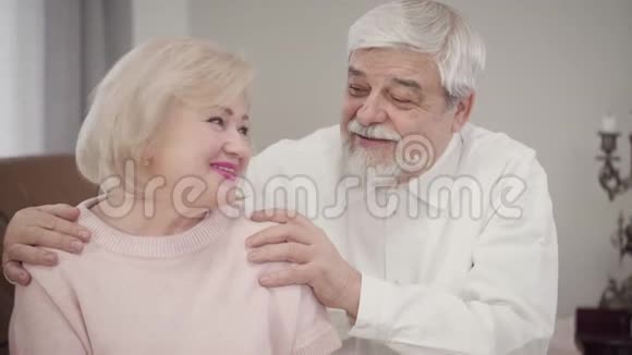年长的高加索夫妇互相交谈看着相机微笑已婚退休人员在室内摆姿势视频的预览图