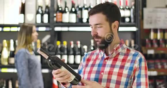 年轻微笑的白人男子在酒瓶上阅读标签的肖像看着相机竖起大拇指富有快乐视频的预览图