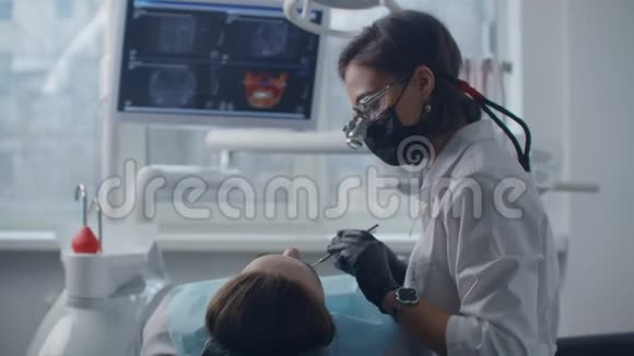 一位年轻的女牙医戴着双筒望远镜和一件白色外套开始检查和治疗一位女性病人牙科治疗视频的预览图