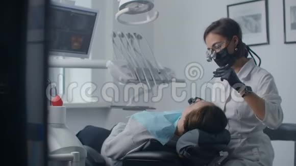 一位年轻的女牙医戴着双筒望远镜和一间白色办公室正在检查和治疗一位女性病人牙科治疗和视频的预览图