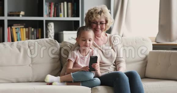 快乐的老奶奶和可爱的孙女在家里使用智能手机视频的预览图