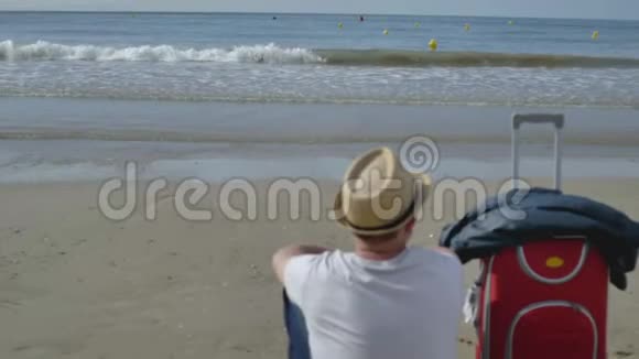 一个戴帽子的年轻人坐在海边的沙滩上旁边是一个红色的手提箱视频的预览图