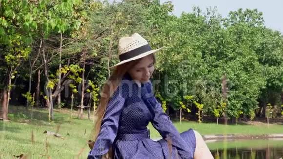 微笑的旅游妇女穿着连衣裙和草帽坐在湖边的草坪上视频的预览图