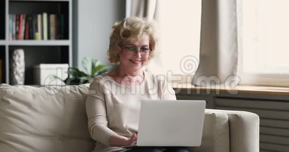 在家里坐在沙发上用笔记本电脑微笑的老女人视频的预览图