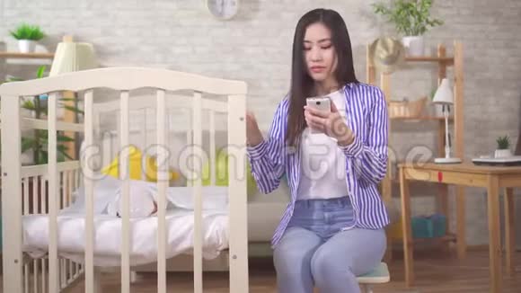 有智能手机的年轻亚洲女人把婴儿抱在婴儿床上视频的预览图