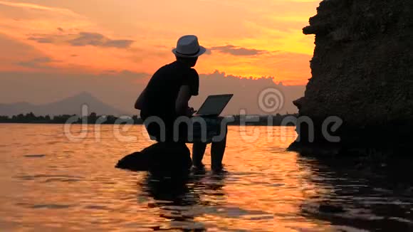 快乐的人坐在石头上在电脑上工作海景静态框架日落海景的男性剪影视频的预览图