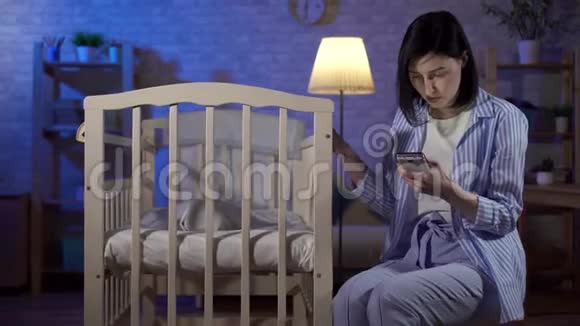 单身年轻母亲在互联网上搜索她手中的智能手机和新生婴儿床视频的预览图