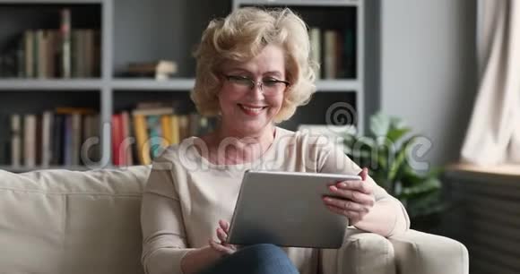 快乐的成熟女人坐在沙发上用数码平板电脑视频的预览图