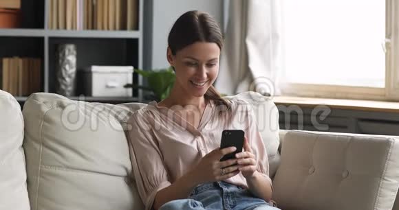 在沙发上用智能手机应用程序微笑迷人的年轻女性视频的预览图