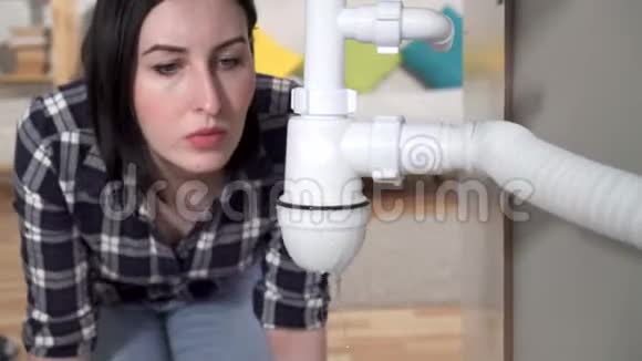穿衬衫的年轻女子发现水槽下面有一个漏水的地方打电话给水管工视频的预览图