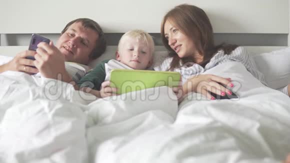 艾米莉和一个儿子早上躺在床上用手机和平板电脑视频的预览图