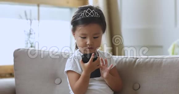 好奇的亚洲小女孩独自坐在沙发上用智能手机视频的预览图