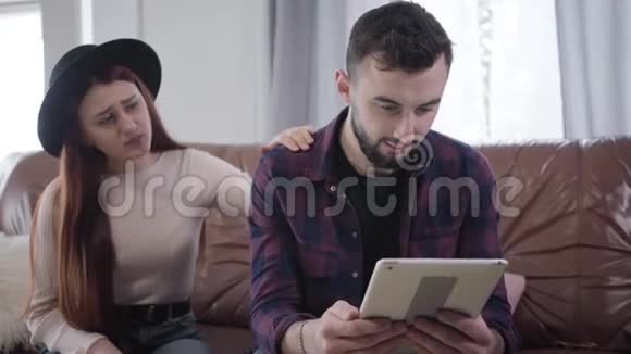 年轻的高加索男人用平板电脑作为他美丽悲伤的女朋友或妻子在后台与他交谈丈夫或丈夫视频的预览图
