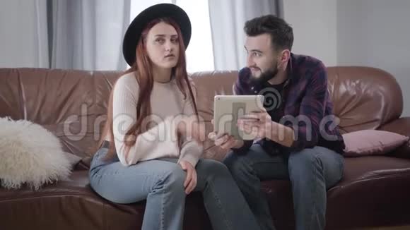 白种人年轻夫妇坐在沙发上用平板电脑说话的肖像穿着优雅帽子签名和视频的预览图