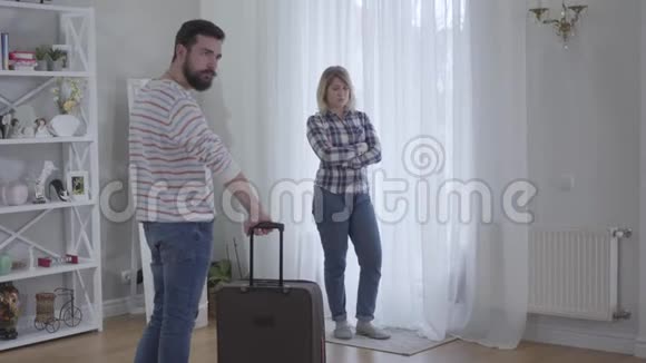 已婚白人夫妇站在室内愤怒地看着对方年轻人拿着旅行袋女人视频的预览图