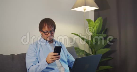 快乐的年轻人使用笔记本电脑和智能手机工作而坐在沙发在家里视频的预览图