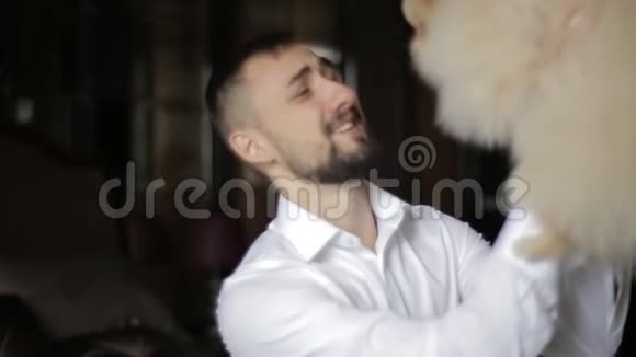 一位身穿白色衬衫的长胡子善良的白种人新郎把心爱的狗抱在怀里在一个房间里吻她视频的预览图