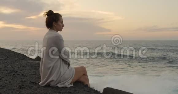 浪漫的白色欧洲女孩坐在巨大的岩石上海滩上日落时的海潮视频的预览图
