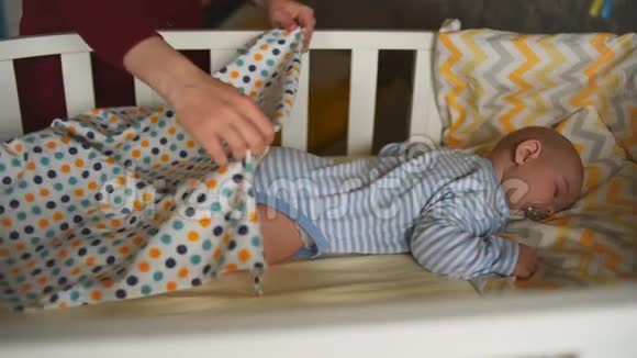 一个小孩子正睡在婴儿床上妈妈用毯子给婴儿盖上被子视频的预览图