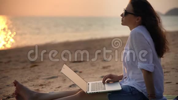 一位年轻的女作家拿着笔记本电脑坐在海滩上打字视频的预览图