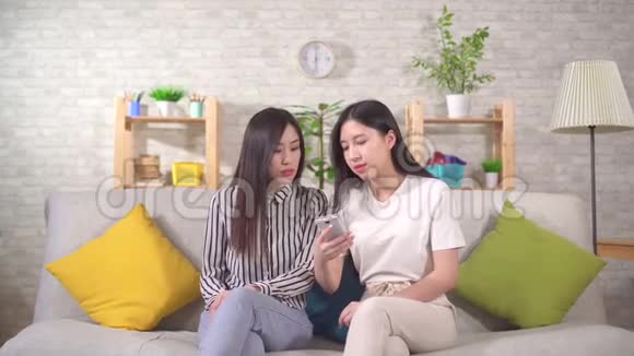 两个快乐的年轻亚洲女人坐在沙发上用智能手机视频的预览图