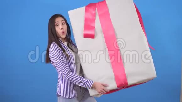 穿着衬衫的积极的亚洲年轻女性在蓝色背景的盒子里拿着一份巨大的礼物视频的预览图
