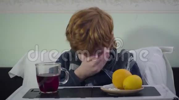 红头发的白人男孩打喷嚏看着摄像机生病的红头孩子坐在家里的床上保健疾病视频的预览图