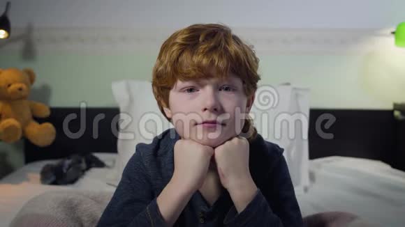 可爱的白种人红头发小孩坐在床上看着摄像机的头像小男孩在卧室里的画像视频的预览图