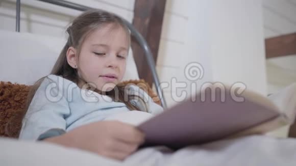 美丽的白种人女孩灰色的眼睛拿着书和阅读躺在床上的小女士的肖像爱好快乐视频的预览图