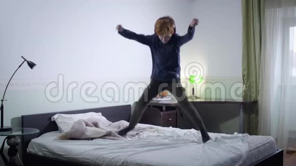 开朗的白人红头发男孩戴着耳机在床上跳跃并有感情地打手势快乐的小孩掉下来视频的预览图