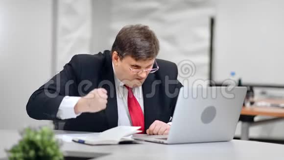 愤怒的男老板愤怒地拍桌子看着红色相机上的笔记本电脑中的屏幕视频的预览图