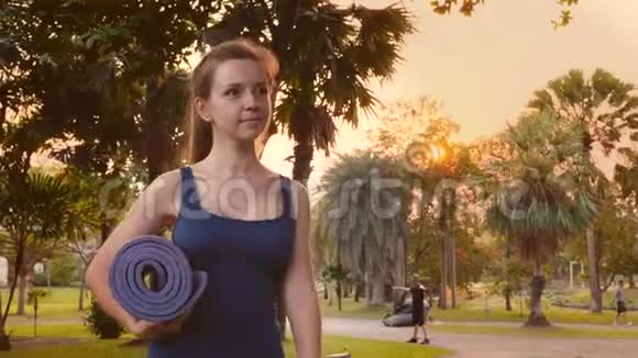 穿着运动装的年轻健身妇女在城市热带公园里用瑜伽垫散步视频的预览图